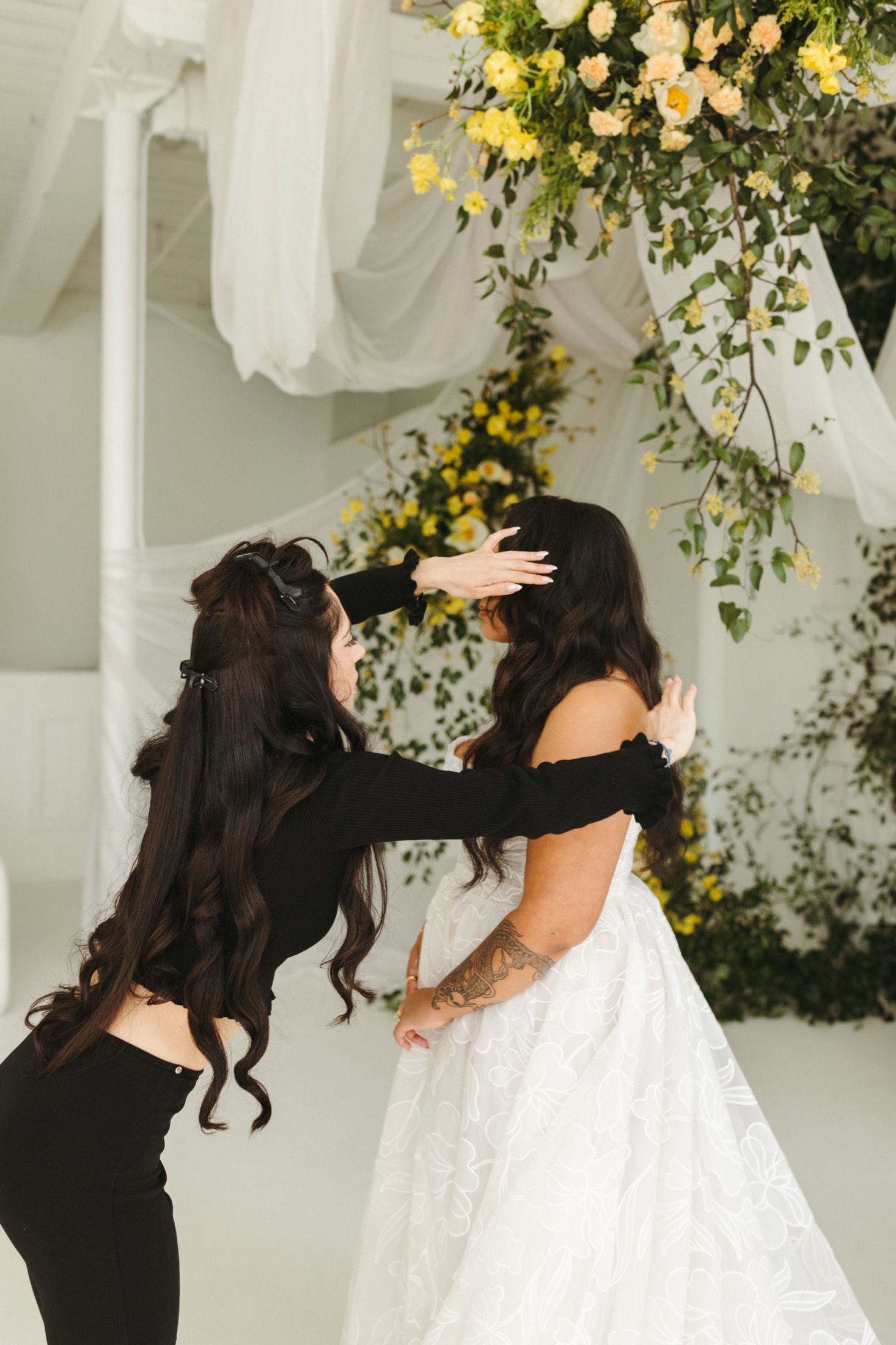 Varvara styling a bride's hair beneath a floral installation before her wedding ceremony